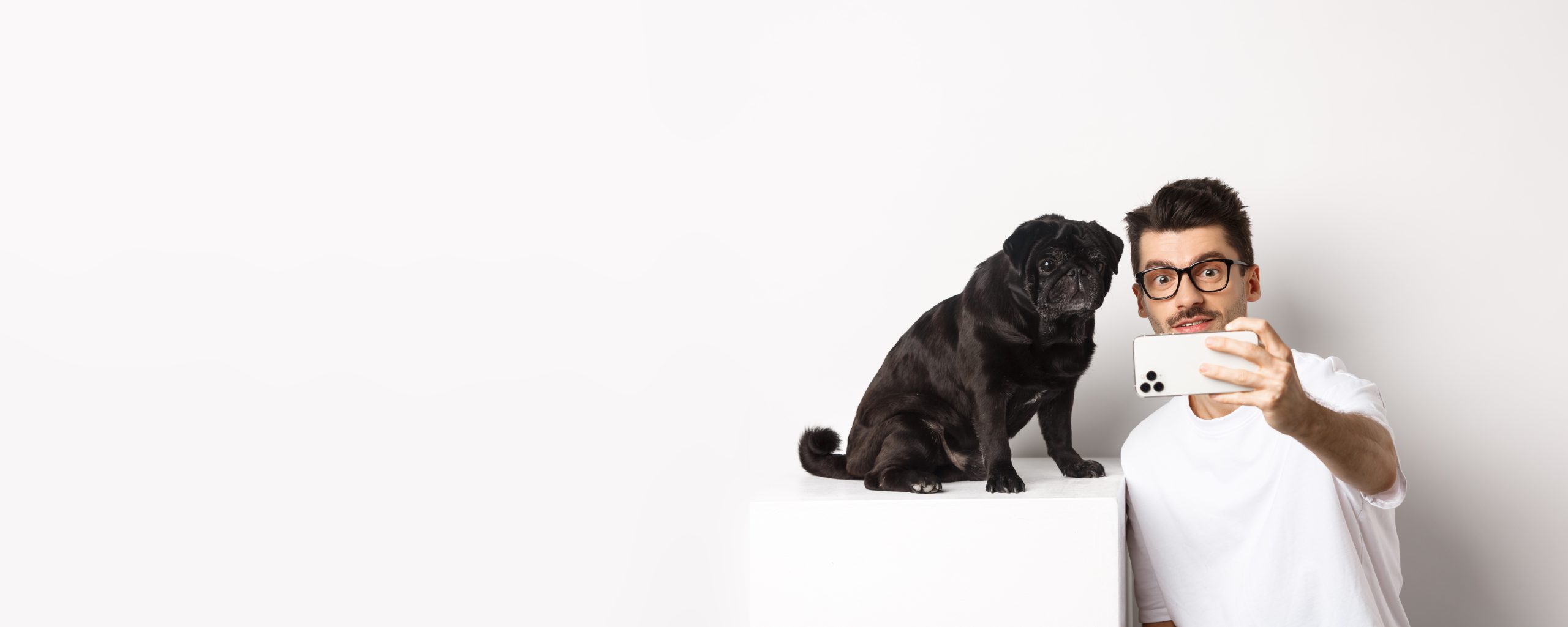 Image of handsome young man taking selfie with cute black dog on smartphone, posing with pug over white background